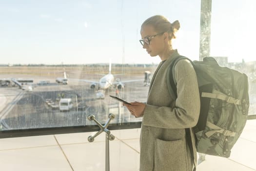 Casually dressed female traveler at airport looking at smart phone device in front of airport gate windows overlooking planes on airport runway.