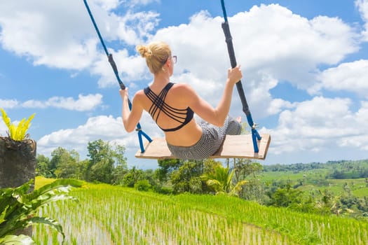 Happy sporty female traveller swinging on wooden swing, enjoying summer vacation in pristine green nature at Jatiluwih rice terraces on Bali, Indonesia. Freedom and life enjoying concept.