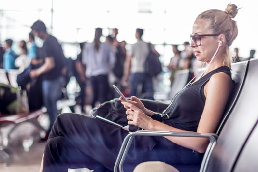 Casual sporty young blond female traveler using her cell phone while waiting to board a plane at the departure gates at the asian airport terminal.