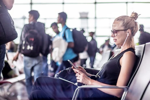 Casual sporty young blond female traveler using her cell phone while waiting to board a plane at the departure gates at the asian airport terminal.