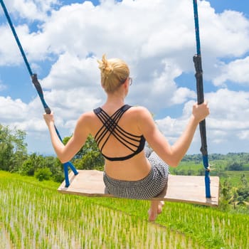 Happy sporty female traveller swinging on wooden swing, enjoying summer vacation in pristine green nature at Jatiluwih rice terraces on Bali, Indonesia. Freedom and life enjoying concept.
