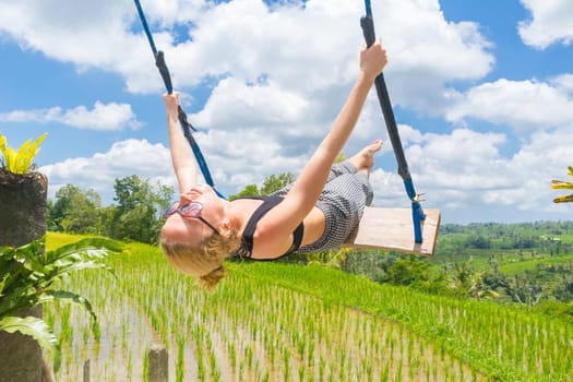 Happy sporty female traveller swinging on wooden swing, enjoying summer vacation in pristine green nature at Jatiluwih rice terraces on Bali, Indonesia. Freedom and life enjoying concept.
