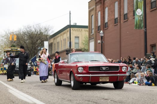 Holland, Michigan, USA - May 12, 2018 A red mustang goes down the road while people dress up with ducth traditional clothing go down the road at the Muziek Parade, during the Tulip Time Festival