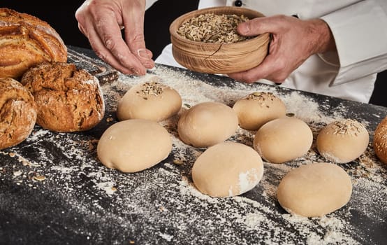 Baker sprinkling crushed grain and seeds on buns dough already formed into buns or rolls on a floured kitchen counter