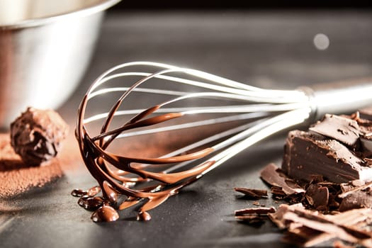 Chocolate coated old vintage metal whisk alongside chopped chocolate and a cacao bonbon on a kitchen table in close up view