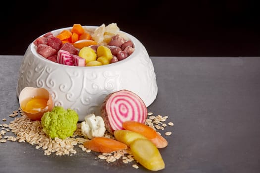 Organic meal for pets mixed in white bowl for cat or dog, consisting of meat, raw egg and fresh vegetables, viewed in close-up against black background