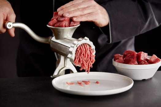 Cook in black uniform mincing fresh beef or pork using old metal manual meat grinder on kitchen counter. Pieces of fresh red meat in white bowl. Cooking preparations, viewed in close-up