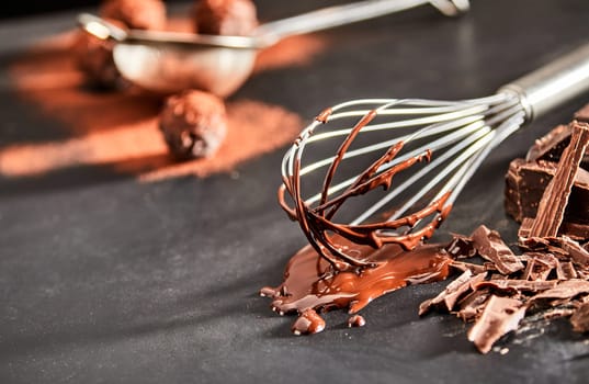 Used metal whisk coated in melted chocolate lying on a kitchen counter with flaked chocolate and copy space