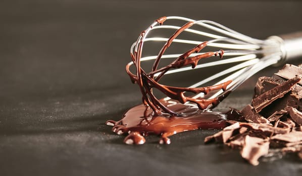 Melted dairy chocolate dripping off an old whisk onto a rustic kitchen table with chopped bar of cooking chocolate alongside