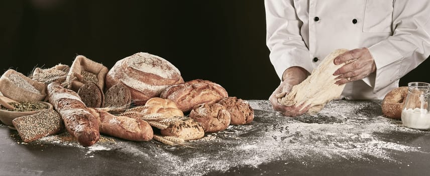 Chef or baker kneading dough while making speciality bread on a black counter with a display of a variety of his products alongside, close up on hands panorama banner