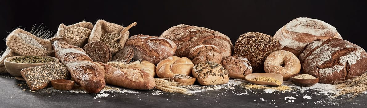 Panoramic banner with rustic bread assortment, decorated with seeds, cereal and flour, sitting on dark surface against white background