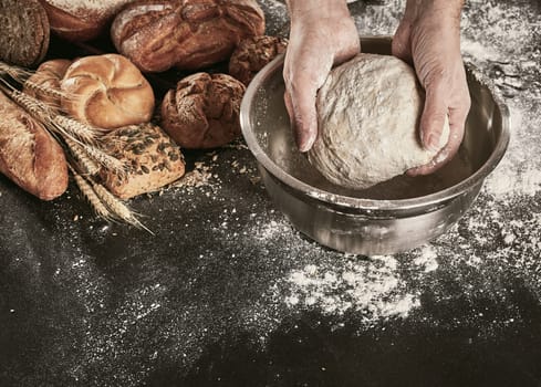 Hands of a baker kneading dough in a metal mixing bowl on a flour covered counter with an assortment of speciality breads alongside