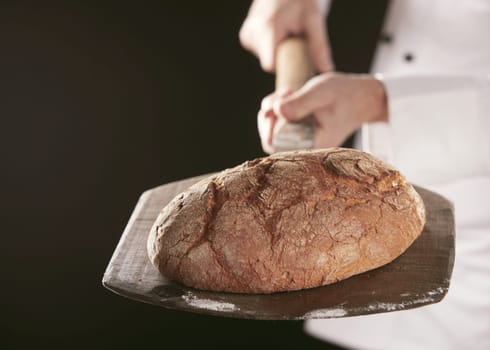 Baker holding loaf of freshly baked bread on wooden shovel, standing in white uniform against black background, viewed in close-up