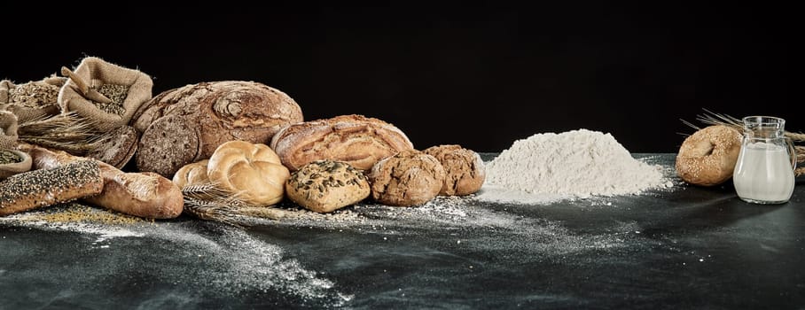 Panorama display of gourmet breads with dough, milk and flour ingredients over a floured kitchen table with copy space