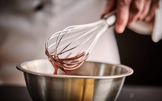 Chef preparing melted chocolate in a mixing bowl using a vintage metal wire whisk in a close up on the utensils