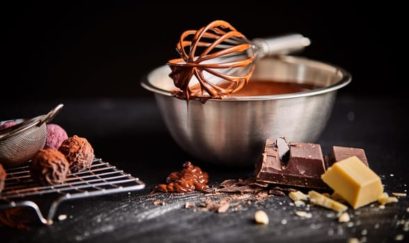Chocolate coated whisk resting on mixing bowl filled with melted chocolate or sauce with a wire rack of bonbons and ingredients