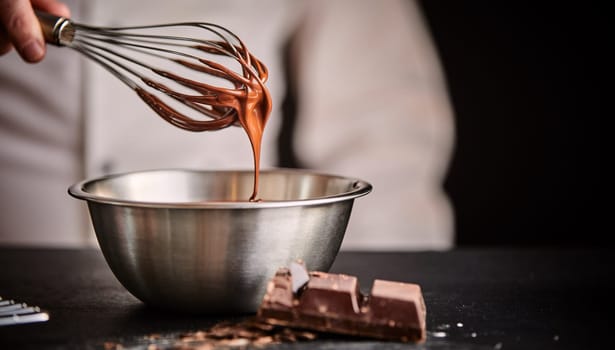 Baker or chef whisking melted chocolate in a bowl with an old metal wire whisk in a close up on his hand and utensils
