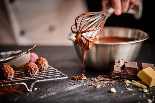 Baker or chocolatier preparing chocolate bonbons whisking the melted chocolate with a whisk dripping onto the counter below