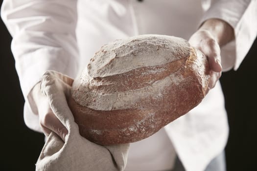 Baker in professional white uniform showing loaf of rye bread in close-up, standing against black background