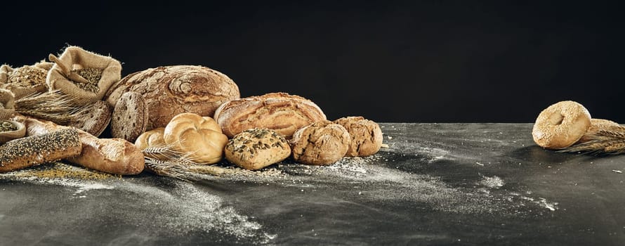 Table with pile of bread assortment. Loaves of white and grey bread, buns and rustic sackcloth bug with cereal, sitting on floured surface against black background, viewed in panoramic banner