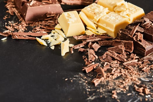 Chocolate for cooking ingredients concept with pieces and shavings of milk and white chocolate, viewed from high angle in close-up on dark surface background with copy space