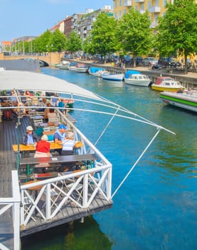COPENHAGEN, DENMARK - JUNE 14, 2018: Sunny summer Copenhagen cityscape with green trees, tourists sitting in restaurant by canal with boats