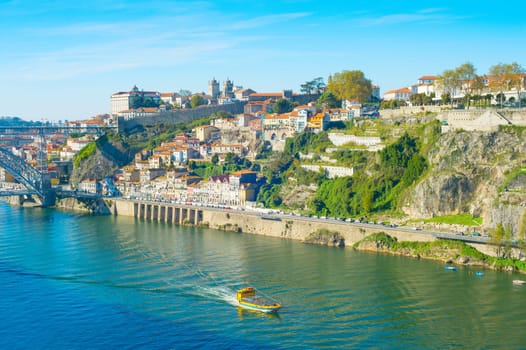 Tourist boat sailing by Douro with Porto cityscape on green riverbank in bright sunlight, Portugal