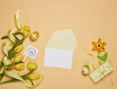 Colored easter eggs, spring tulips flowers, gift box and blank sheet with envelope on yellow background. Top view with copy space. Greeting card, Easter background