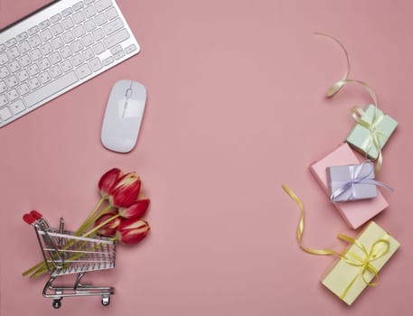 Shopping trolley, gift boxes, tulips flowers, computer keyboard and mouse on pink background. Top view with copy space. Valentine's day, Mother's day, Women's day, wedding. Seasonal sale