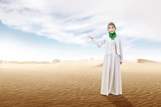 Pretty asian muslim woman with open palm showing something on the sand dune with blue sky background