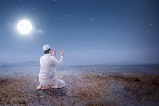 Rear view of asian muslim man sitting in pray position while raised hands and praying on the sand dune with moon and night scene background