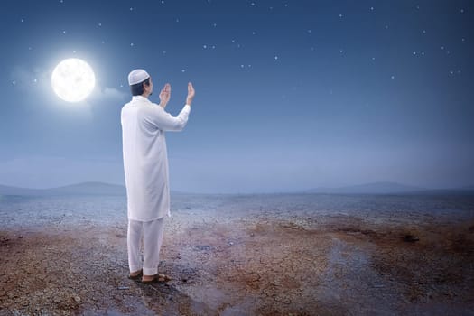 Rear view of asian muslim man raised hands and praying on the sand dune with moon and night scene background