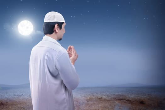 Rear view of asian muslim man raised hands and praying on the sand dune with moon and night scene background