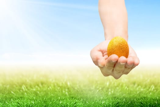 Human hand holding yellow easter egg on the grass field with sunlight and blue sky background. Happy Easter