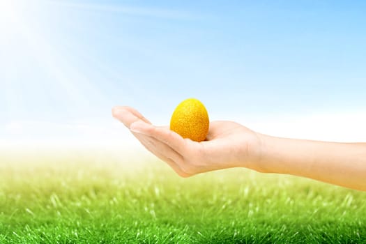 Human hand holding yellow easter egg on the grass field with sunlight and blue sky background. Happy Easter