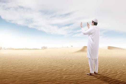 Rear view of asian muslim man raised hands and praying on the sand dune with blue sky background