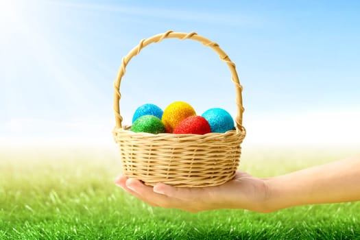 Human hand holding wooden basket with colorful easter eggs on the grass field with sunlight and blue sky background. Happy Easter