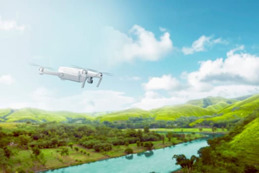 White drone with camera flying over green hills with river and trees over blue sky background