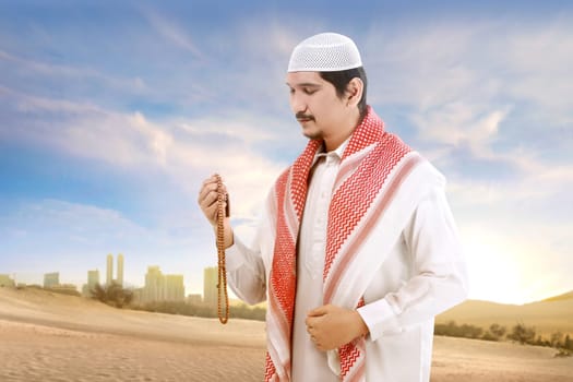 Young asian muslim man with cap and turban standing and praying with prayer beads on the sand with blue sky background