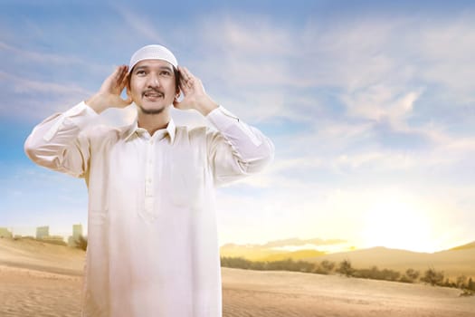Smiling asian muslim man with cap standing and praying on the sand with blue sky background
