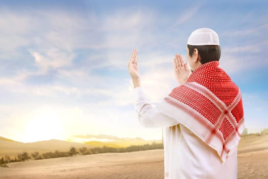 Rear view of asian muslim man with cap and turban standing and praying with raised arms on the sand with blue sky background