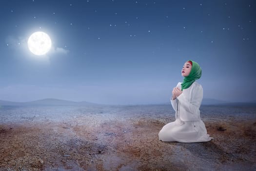 Young asian muslim woman sitting in pray position while raised hands and praying on the sand dune with moon and night scene background