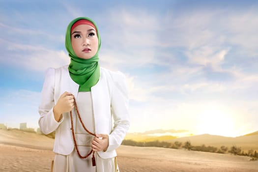 Young asian muslim woman in veil standing and praying with prayer beads on the sand with blue sky background