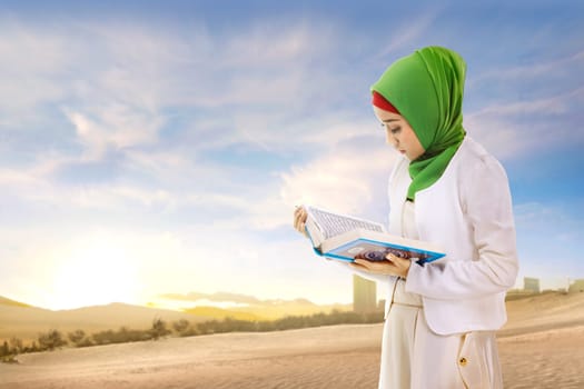 Beautiful asian muslim woman in veil standing and reading the quran on the sand with blue sky background