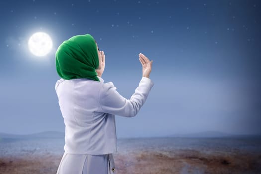 Rear view of asian muslim woman in veil raised hands and praying on the sand dune with moon and night scene background