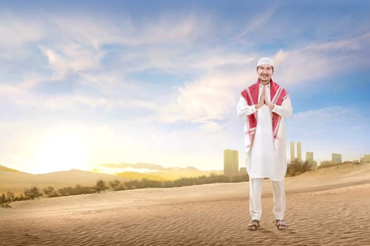 Smiling asian muslim man with cap and turban standing and praying on the sand with blue sky background