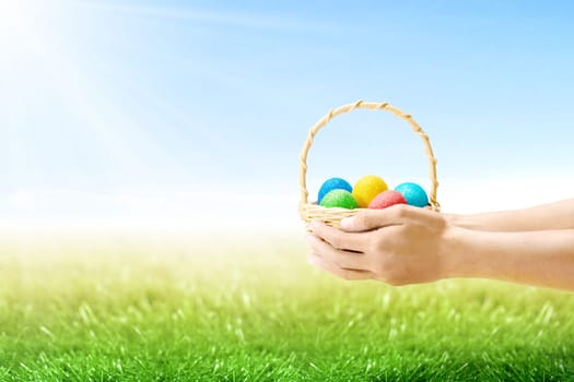 Human hands holding wooden basket with colorful easter eggs on the grass field with sunlight and blue sky background. Happy Easter