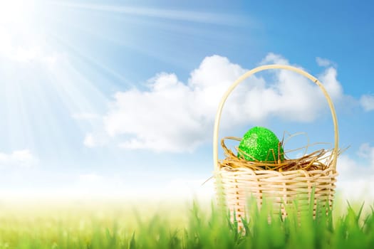 Green easter egg in the nest on wooden basket on the grass field with sunlight and blue sky background. Happy Easter