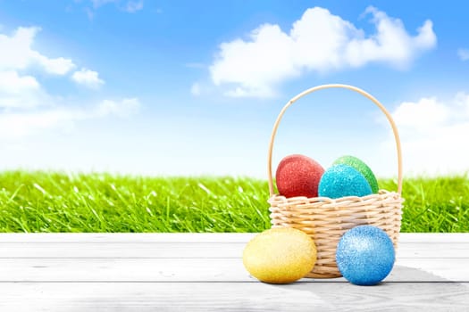 Colorful easter eggs in wooden basket and wooden table with grass field and blue sky background. Happy Easter