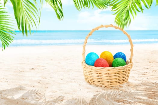 Colorful easter eggs in wooden basket on the sandy beach with palm branch and waving sea over blue sky background. Happy Easter